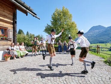 Bauernherbst Schuhplattler Men in Austrian traditional costume dance outside a wooden house, with mountains and people in the background.
