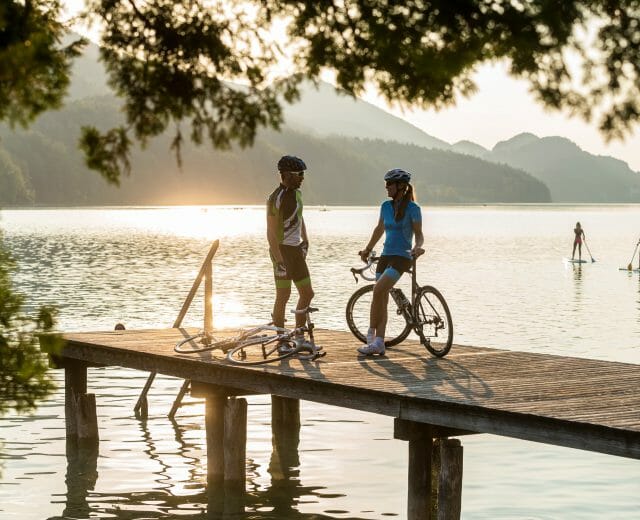Two triathlon cyclists talk on a wooden jetty by a lake at sunset, paddleboarders in the background.