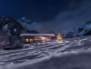 A cosy hut with lights on, surrounded by snow and mountains under a starry night sky.
