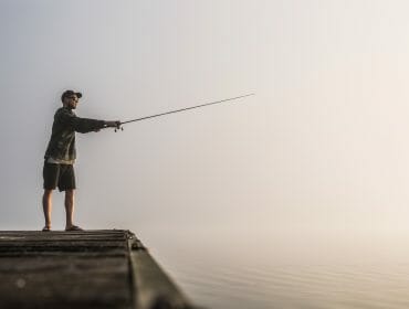 A person takes action, standing on a quay fishing in the early morning fog over calm water.