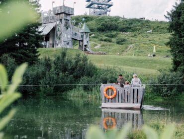 Three people ride a wooden raft across a pond in a green park with a large wooden structure in the background.