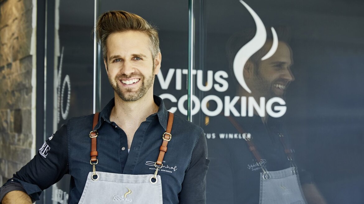 Smiling man in an apron stands in front of a glass door with the Vitus Cooking logo. (Enlarged view)