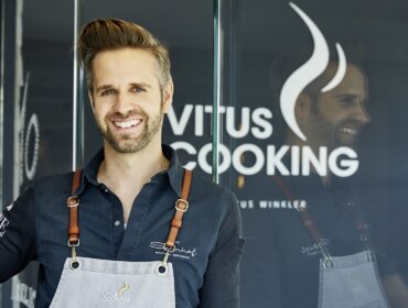 Smiling man in an apron stands in front of a glass door with the Vitus Cooking logo.