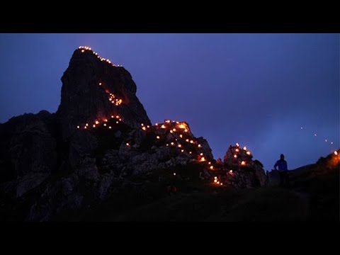 A rocky mountain at dusk with a trail of glowing lights, reminiscent of Alpine summer events.