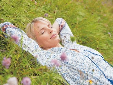 agr_stresspraevention Woman lying in tall green grass with eyes closed, relaxing and smiling, surrounded by wildflowers.