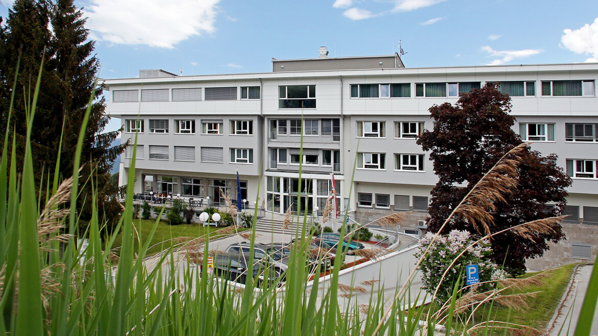 A modern, three-storey building with large windows, surrounded by trees and grass on a sunny day.