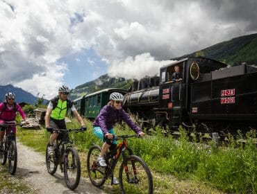 Bahnen im SalzburgerLand Three cyclists ride beside a black steam train on a gravel path in a scenic mountainous area under cloudy skies.