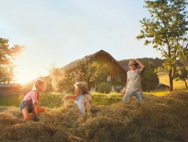Three children play in a haystack near a barn on a sunny day in the countryside.