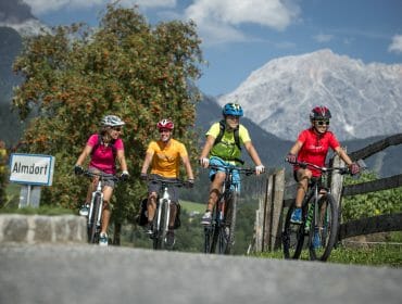 Four people riding bicycles on a country road with mountains and greenery in the background, near an Almdorf sign.