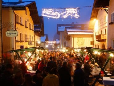 taxenbach-winter-advent Crowds browse festive market stalls in a brightly lit, snowy village street decorated with Christmas lights.