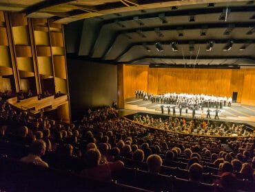 A large audience watches a performance on stage in a modern theatre with an orchestra in the pit.