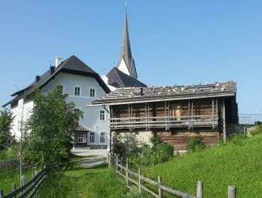 A white church with a tall steeple and a wooden barn sit on a grassy hill under a clear blue sky.