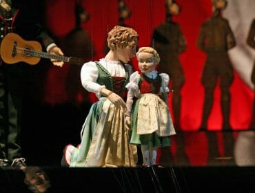 Two marionette puppets in traditional attire perform on stage with a guitar player and shadowy figures behind them.