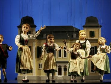 Marionette puppets dressed in österreichisch traditional costumes perform in front of a house backdrop on stage.