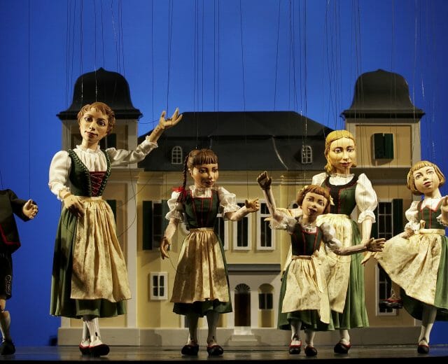 marionettentheater sound of music Marionette puppets dressed in österreichisch traditional costumes perform in front of a house backdrop on stage.