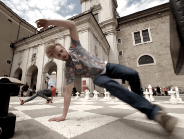 A man breakdancing on a large outdoor chessboard in front of historic buildings.