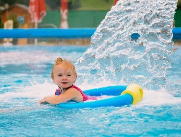 Smiling toddler in a swimming pool floats on a blue rubber ring under a water fountain at thermal baths for families.
