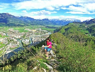 Blick-vom-Barmstein-auf-Hal Person sitting on a hilltop overlooking a river, town, and mountains under a partly cloudy sky.