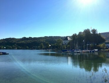 A calm lake with sailboats by the shore, trees, and buildings under a clear blue sky with bright sunlight.