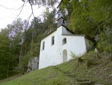 Falkenstein00010 Small white chapel with a bell tower sits on a grassy hillside, surrounded by dense green forest.