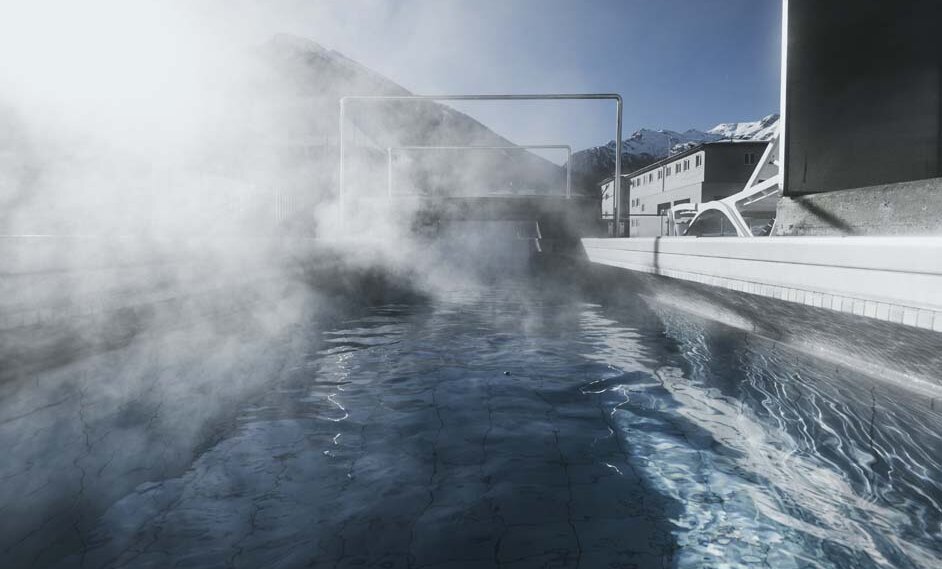 Outdoor hot pool with steam rising, next to a sauna, surrounded by buildings and snow-capped mountains in the background.
