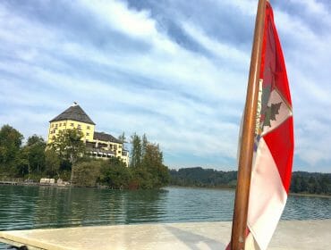 A red and white flag by a lake with a castle-like building and trees in the background under a blue sky.