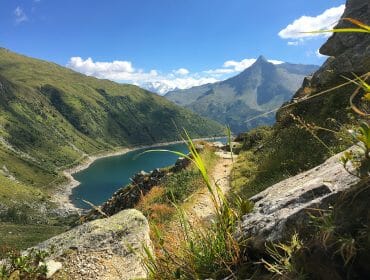 Mountain trail overlooking a blue lake, surrounded by green hills and distant peaks under a blue sky.