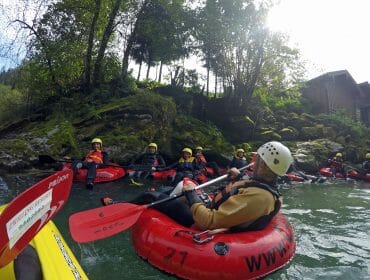 People in helmets and wetsuits float on red lilos in a river near a forested, rocky shore.