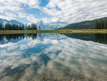 A clear lake reflects clouds, trees, and distant mountains—perfect for 4 Lakes Art Hike inspiration.