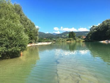 Calm river with sandy banks, trees, distant hills, and people relaxing under a clear blue sky.