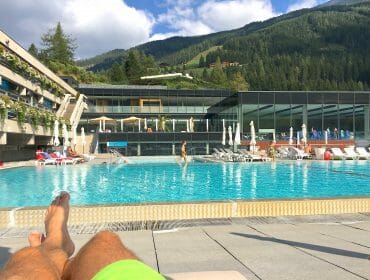 Person relaxing by an outdoor swimming pool with modern buildings and mountains in the background.