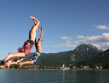Two boys in swimming costumes jump off a jetty into a lake, with mountains and blue sky in the background.