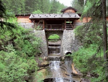 A rustic stone and wood bridge, perfect for trifting, spans a small waterfall in a lush green forest.