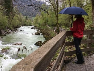 Hiking in the rain, a person in a red jacket with a blue umbrella stands on a wooden bridge by a forest stream.
