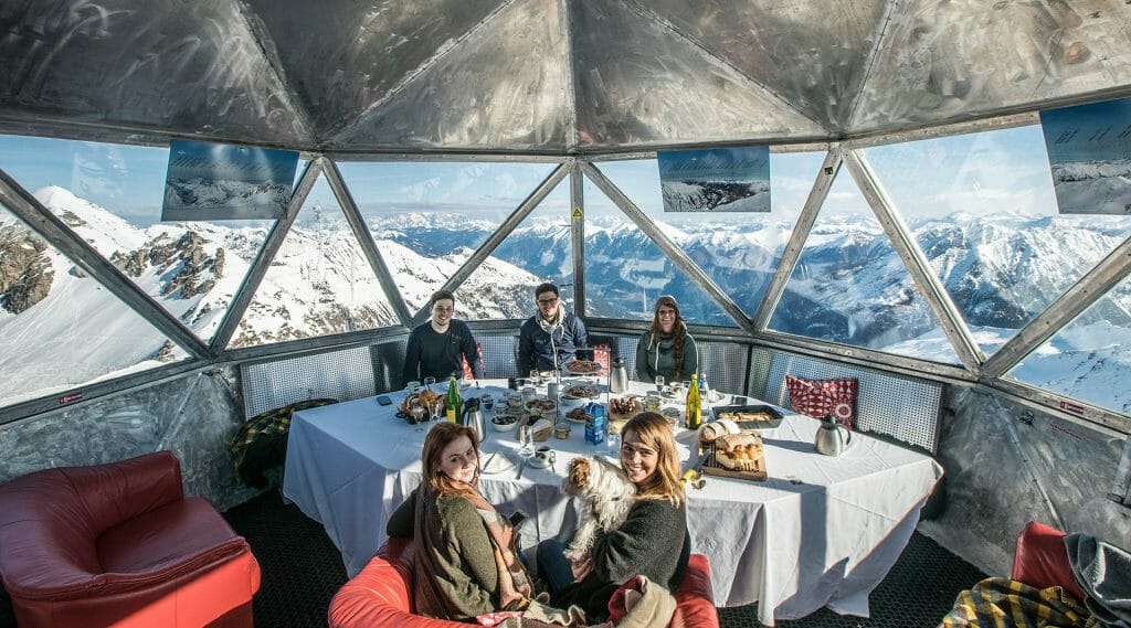 a group of people eating on a metal building with snowy mountain in the background