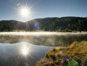 04-Herbststimmung-am-Preber Bright sun over Lungau’s misty lake, sunlight reflecting in calm water and grassy shore in front.