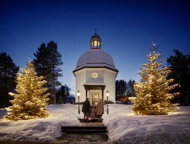 Two people enter a snowy chapel at dusk, flanked by lit Christmas trees and glowing lamps.