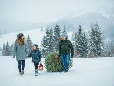 A family walks in the snow, carrying a Christmas tree and lantern, enjoying festive Christmas events.