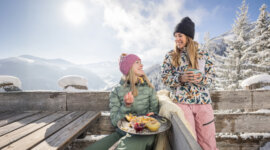 chillout-huettengenuss2-1024×569 Two women in winter clothing enjoy food and drinks outdoors, with snow-covered mountains and trees in the background.