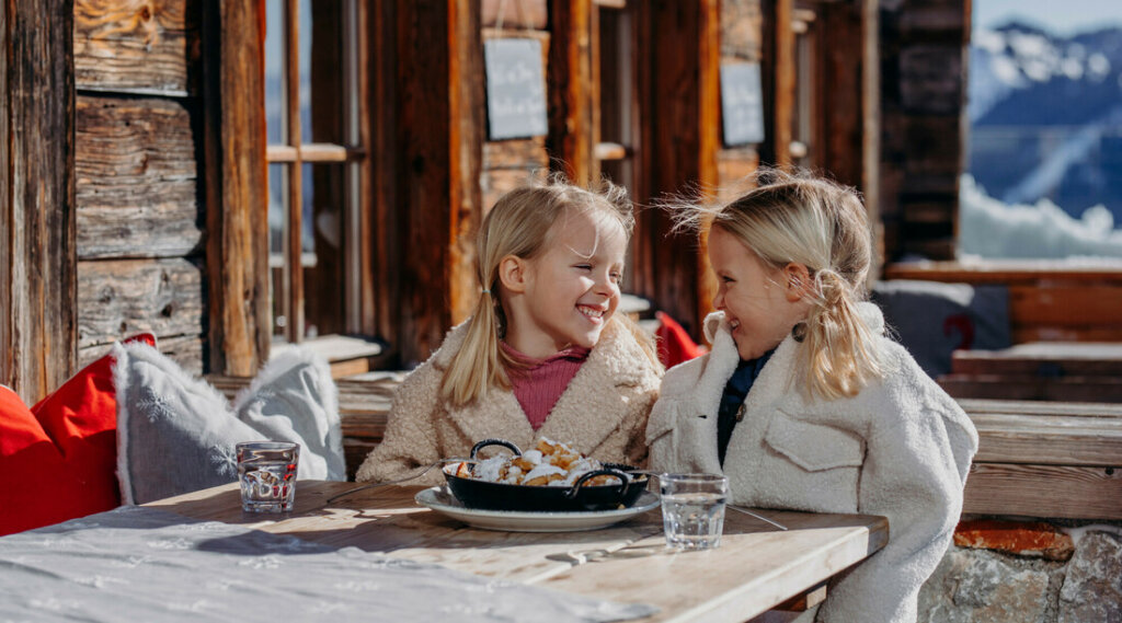 chillout-kinder-gaumenfreu © SalzburgerLand Tourismus, Verena Schierlden Two young girls in warm coats smile at each other whilst sitting outside at a table with a plate of food. (Enlarged view)