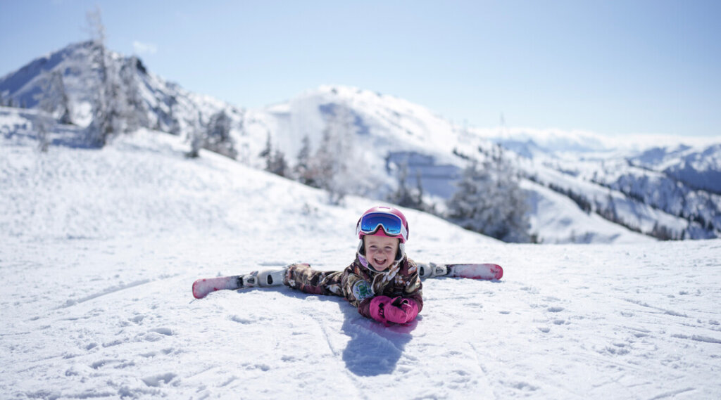 Smiling child in ski gear does the splits on snowy slope with mountains in the background.