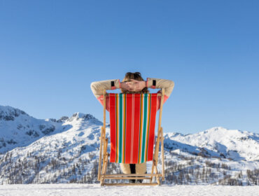 Person relaxing in a striped deckchair, facing snow-covered mountains under a clear blue sky.