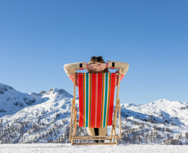 Person relaxing in a striped deckchair, facing snow-covered mountains under a clear blue sky.