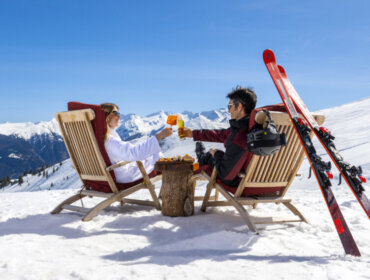 Two people in ski gear toast drinks whilst relaxing in chairs on a snowy mountain with skis nearby.