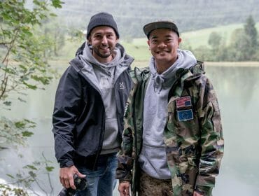 Two men in jackets stand smiling by a lake, with trees, mountains, and Kenneth Nguyen in the background.