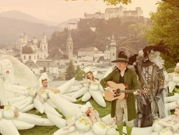 People in whimsical costumes, including flowers, angels, and fantasy figures, gather on a grassy hill above a city.