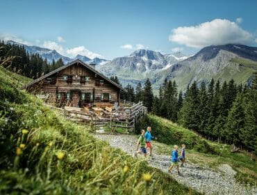 Four children hike a mountain trail past a wooden cabin with alpine scenery and tall pine trees in the background.