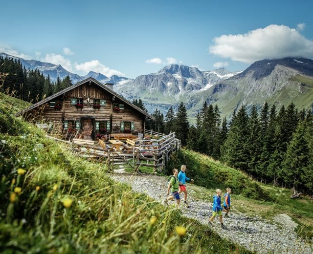 Four children hike a mountain trail past a wooden cabin with alpine scenery and tall pine trees in the background.