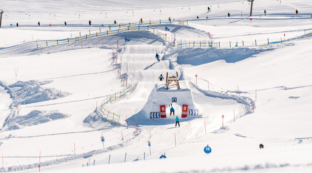 (c) christian-riefenberg Skiers descend a snowy terrain park with jumps and gates on a clear winter day. (Enlarged view)