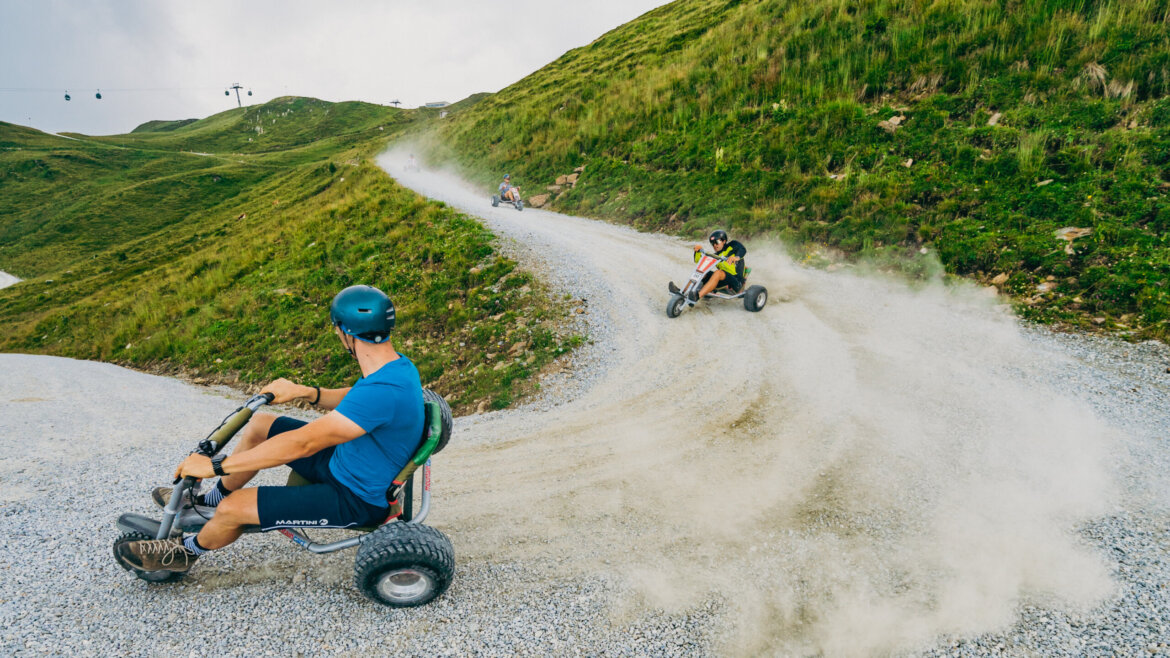(c) Wildkogel Arena People riding drift trikes on a winding gravel mountain track, kicking up dust as they turn. (Enlarged view)